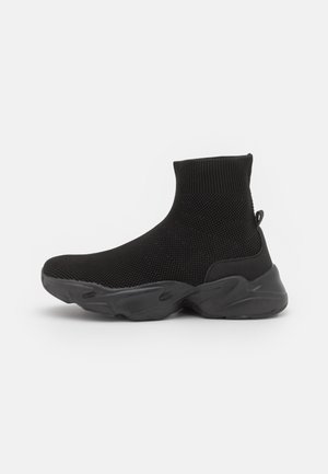 Basket type chaussette en maille noire avec semelle épaisse sculptée et languette sur fond uni gris clair.