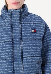Blauwe denim pufferjack met een gewatteerde textuur, voorzien van een logo-print over de gehele oppervlakte en een klein logopatch op de linkerborst. Hoog kraagontwerp.