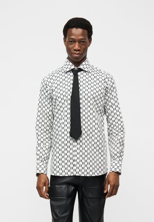 Mężczyzna w białej koszuli z czarnym geometrycznym wzorem, czarnym krawatem i czarnych skórzanych spodniach, stojący na tle jednolitego jasnego tła.