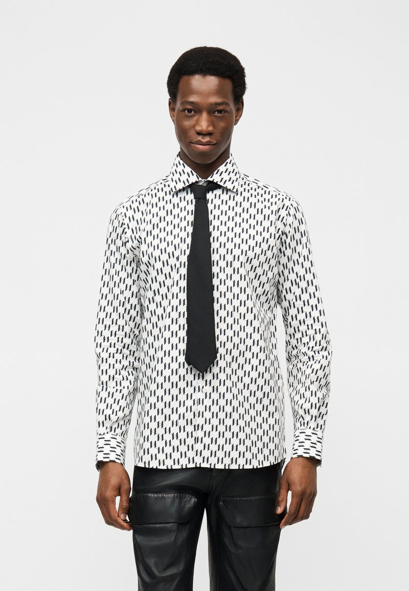 Άντρας φοράει λευκό πουκάμισο με μαύρο γεωμετρικό σχέδιο, μαύρη γραβάτα και μαύρο δερμάτινο παντελόνι, στέκεται μπροστά σε απλό φωτεινό φόντο.