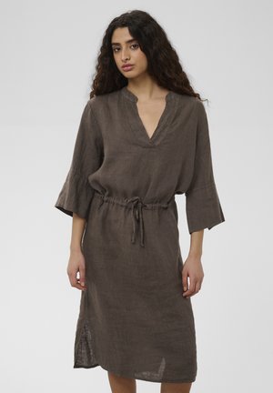 Woman with curly hair wearing a loose, knee-length, brown dress with three-quarter sleeves and a waist drawstring, standing against a plain background.