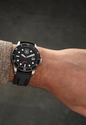 Montre-bracelet avec cadran noir, lunette tournante, guichet date et bracelet en caoutchouc noir portée au poignet gauche sur fond sombre.