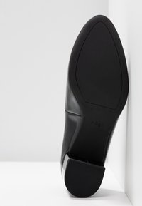 Chaussure à talon haut en cuir verni noir avec un extérieur lisse et un bout arrondi. La semelle est en caoutchouc noir avec une texture rainurée horizontale.