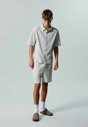 Chemise à manches courtes gris clair et shorts assortis avec un motif texturé, associés à des slippers marron et des chaussettes blanches jusqu'à la cheville.