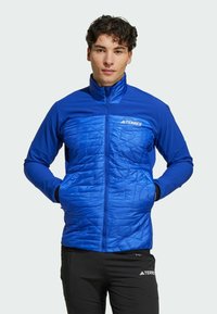 Blauwe geïsoleerde jas met een hoge kraag, gestructureerde voorkant, ritszakken en een logo op de borst, gecombineerd met zwarte broek.