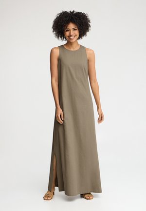 Uśmiechnięta kobieta o kręconych włosach, ubrana w oliwkowozieloną, bezrękawnikową sukienkę maxi z rozcięciem po boku oraz brązowe sandały na otwartym palcu, stoi na tle białego tła.