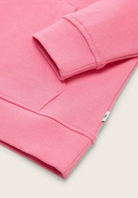 Felpa rosa con polsini e orlo a coste, texture morbida e dettaglio cucito diagonale vicino allo scollo. Piccola etichetta del marchio visibile.