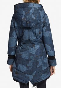Kvinde iført lang, blå camouflage-quilted jakke med hætte og justerbare snore, vist fra ryggen.