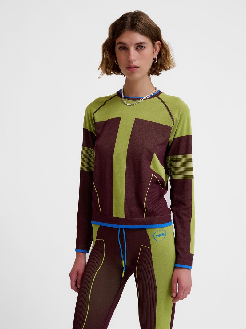 Jeune personne portant un haut à manches longues et des leggings assortis verts et marron foncé à motifs avec un liseré bleu et un collier, debout devant un fond blanc.