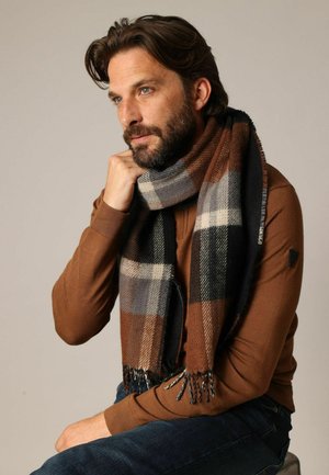 Écharpe à carreaux dans des tons de brun, gris et noir, en tissu doux avec des bords effilochés. Le modèle la porte drapée sur un pull marron.