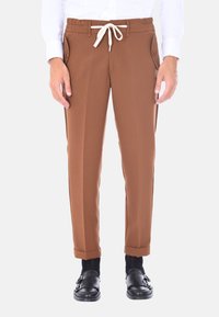 Pantaloni marroni a gamba affusolata realizzati in tessuto liscio, con vita regolabile tramite cordoncino e tasche laterali con chiusura a bottone. Cuffs arrotolati sopra le scarpe.