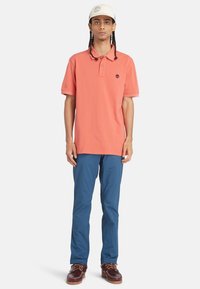 Perzikkleurige polo met kraag en logo, gecombineerd met blauwe broek en bruine schoenen. De outfit heeft een casual, ontspannen pasvorm.