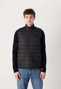 Colete puffer preto com design acolchoado vertical, gola alta e fecho de correr frontal, combinado com uma camisa preta de manga longa e uns jeans azuis.