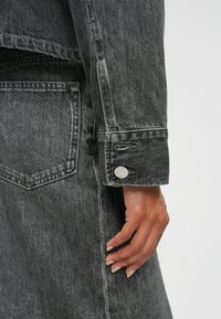 Czarna dżinsowa kurtka z wyraźnie zarysowanym kołnierzem, widoczną kieszenią i okrągłym metalowym guzikiem na mankiecie. Tkanina ma wyblakłą teksturę.