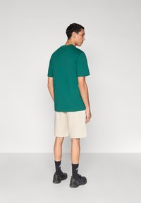 Πράσινο βαμβακερό T-shirt με κοντά μανίκια, φορεμένο με μπεζ σορτς. Ορατά μαύρα αθλητικά παπούτσια και κάλτσες, σε ουδέτερο φόντο.