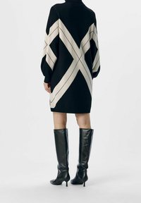 Zwart en crèmekleurig sweaterjurk met een gedurfde diagonale streep patroon, voorzien van een hoge hals en ballonmouwen, gecombineerd met glanzende zwarte kniehoge laarzen.