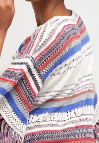 Poncho in maglia con strisce rosse, blu e bianche, dettagli in pizzo e frange. Tessuto leggero e strutturato.