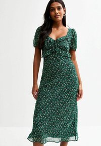 Robe midi florale verte avec des manches courtes bouffantes, un détail à nouer au niveau de la poitrine et un corsage ajusté. Tissu léger et fluide avec une finition semi-transparente.