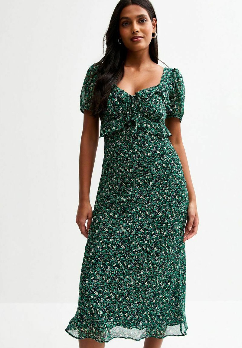 Robe midi florale verte avec des manches courtes bouffantes, un détail à nouer au niveau de la poitrine et un corsage ajusté. Tissu léger et fluide avec une finition semi-transparente.