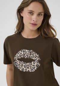 Hnědé bavlněné tričko s grafikou rtů se leopardím vzorem v krémové a černé barvě. Kulatý výstřih a krátké rukávy. Hladká textura.