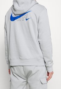 Grå hoodie med stor blå Nike-logotyp på baksidan. Har huva med dragsko och ribbade muddar. Materialet verkar mjukt och strukturerat.