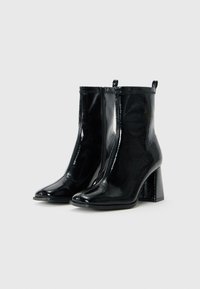 Une paire de bottines en cuir verni noir brillant avec des bouts carrés, des talons carrés, des zips sur le côté et des languettes de tirage sur un fond blanc.