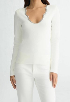 Vrouw draagt een wit top met lange mouwen en een gegolfde halslijn afgezet met zilveren strass-steentjes en witte broek.