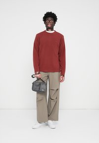 Chemise rouge à manches longues, pantalon beige à jambes larges, baskets blanches, tenant un sac gris avec logo. Design simple, sans motifs, texture lisse.