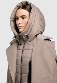 Doudoune beige avec un grand col à capuche. Dispose d'une fermeture éclair à l'avant, d'une texture matelassée et d'un tissu bicolore avec des sections mates et brillantes.