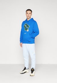 Fleece azul com o logótipo "Bucks", bolso canguru e cordão. Combinado com calças azul claro e ténis preto e branco.