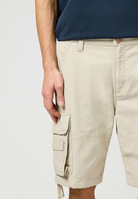 Beiga cargoshorts med flera lapplommar med klaffstängning, en slät tygtextur och justerbara dragsnören vid nederkanten.