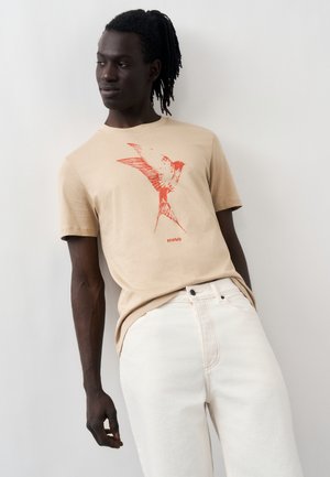 Homme à la peau foncée et aux dreadlocks portant un T-shirt beige avec un motif d'oiseau rouge et un pantalon blanc, appuyé légèrement contre un mur blanc.