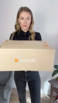 Scatola di cartone rettangolare marrone con la scritta "zalando" stampata in bianco. Sostenuta da una persona che indossa un top nero e jeans scuri, con una pianta verde nelle vicinanze.