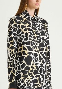 Woman wearing black and beige giraffe-print long-sleeve button-up shirt with gold hoop earrings, on a plain light background.