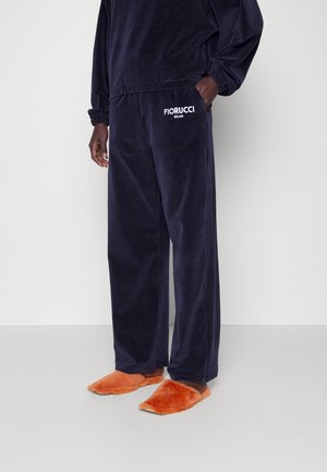 Osoba ubrana w ciemnogranatowe welurowe spodnie Fiorucci i dopasowany top, w pomarańczowych futrzastych kapciach, stojąca na jednolitym jasnym tle.