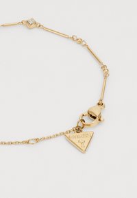Bracelet en ton or présentant des maillons allongés et ronds, une pierre carrée transparente et un pendentif triangulaire avec "GUESS" en relief. Fermeture à coulisse.
