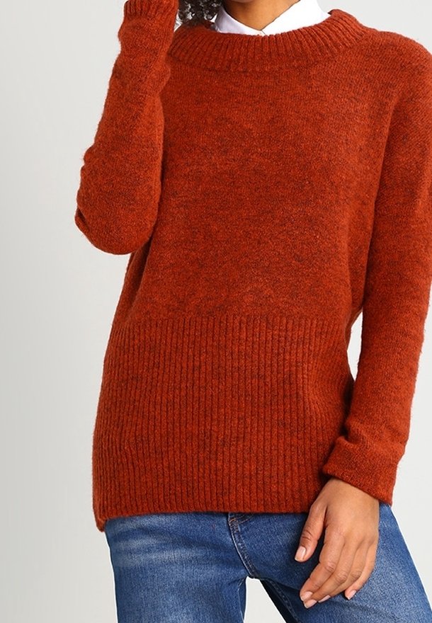 Femme portant un pull en tricot couleur rouille avec des détails côtelés, associé à un jean en denim bleu, posant avec une main près du visage.