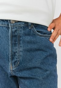 Jean en denim bleu foncé avec une texture lisse, présentant un zip et une fermeture à bouton, des coutures visibles et des poches avant.
