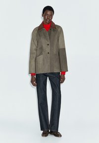 Persona che indossa un giubbotto bicolore marrone, un maglione a collo alto rosso, pantaloni jeans neri dritti e scarpe marroni, in piedi contro uno sfondo semplice.