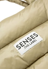 Beige gepolsterte Jacke mit glänzender Oberfläche und horizontalen Nähten. Verfügt über ein rechteckiges Etikett mit "SENSES THE LABEL" in Schwarz darauf.