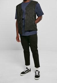 Černá prošívaná vesta s předním zipem, nošená přes tmavě modré tričko, spárovaná s černými kalhotami a černými teniskami s bílými akcenty.