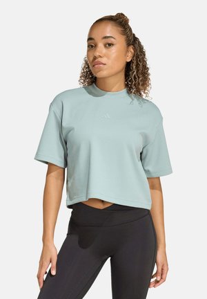 Joven mujer con cabello rizado que lleva una camiseta corta azul claro y leggings negros de cintura alta, de pie frente a un fondo liso.