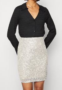 Czarna, teksturowana koszula na guziki z falbaniastym kołnierzem oraz dopasowana mini spódniczka z srebrnych cekinów. Kontrast materiałów i blasku przyciąga uwagę wizualną.