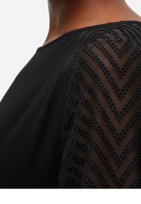 Blusa negra con mangas transparentes con patrón de chevron y detalles texturizados. Escote redondeado y tejido liso en el cuerpo.