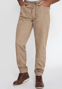 Beige, lichtgewicht broeken met een elastische tailleband, een trekkoord en zijzakken. Tapered pijpen met cuffs aan de onderkant. G gedragen met bruin schoeisel.