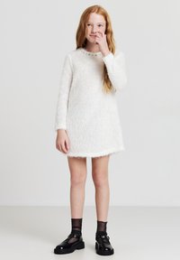 Abito bianco e soffice con maniche lunghe, scollatura rotonda e una finitura testurizzata. Presenta bottoni decorativi all'uscita del collo e una vestibilità ampia.