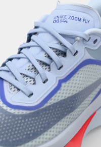 Zapatillas Nike Zoom Fly de color azul claro con parte superior de malla, acentos grises, detalles púrpuras y suela texturizada. Cintas y logotipo visibles.