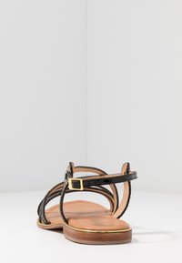 Sandalo nero con cinturini, caratterizzato da una parte superiore testurizzata, dettaglio della fibbia dorata e una suola in legno marrone chiaro. Design semplice con schienale aperto.