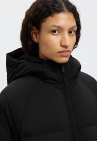 Zwarte donsjas met een hoge kraag en capuchon. Voorzien van een ritssluiting en gewatteerde structuur. Minimalistisch ontwerp, gladde stof.