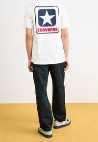 White cotton t-shirt with a blue square and white star logo on the back. Paired with dark pants and multi-colored sneakers.